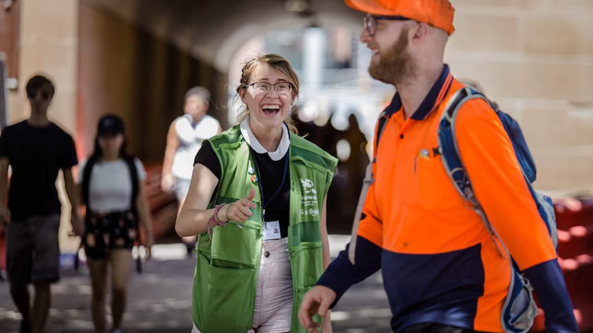 A smiling charity volunteer in a green vest giving a thumbs up to a passer-by in a high-vis shirt