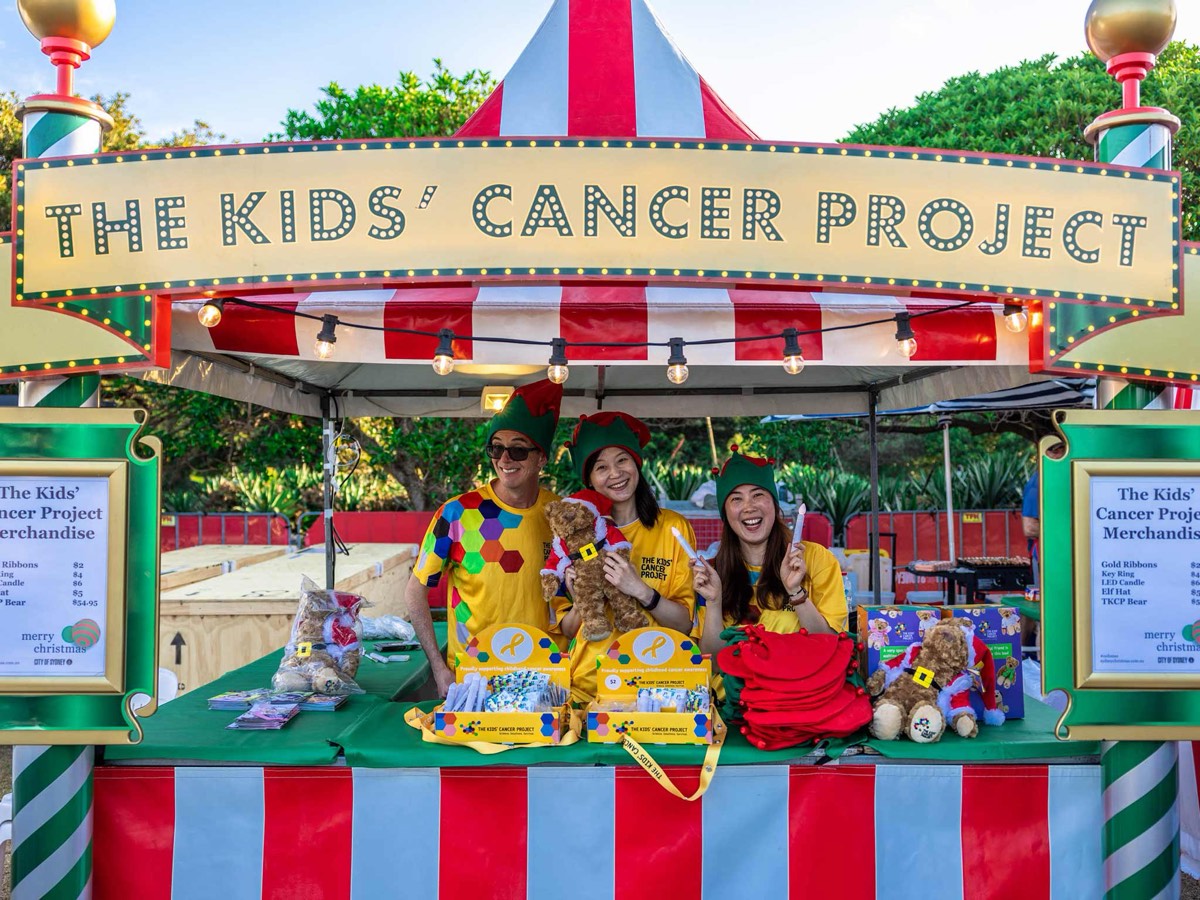 Three volunteers in elf hats and yellow Kids' Cancer Project t-shirts at a red-and-white striped Christmas market stall, holding up teddy bears and LED candles