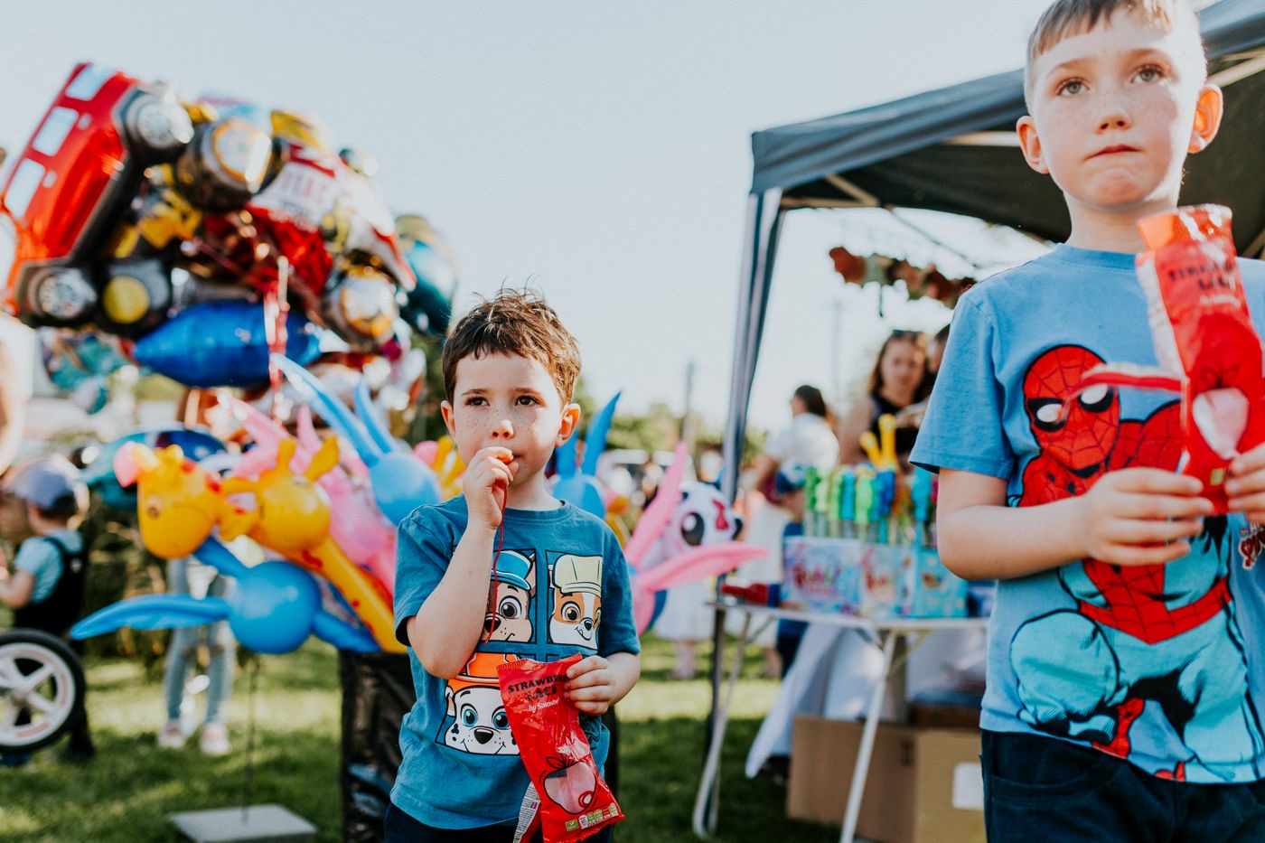 Two boys eating ice blocks at a school fete stall, balloons and a marquee behind them